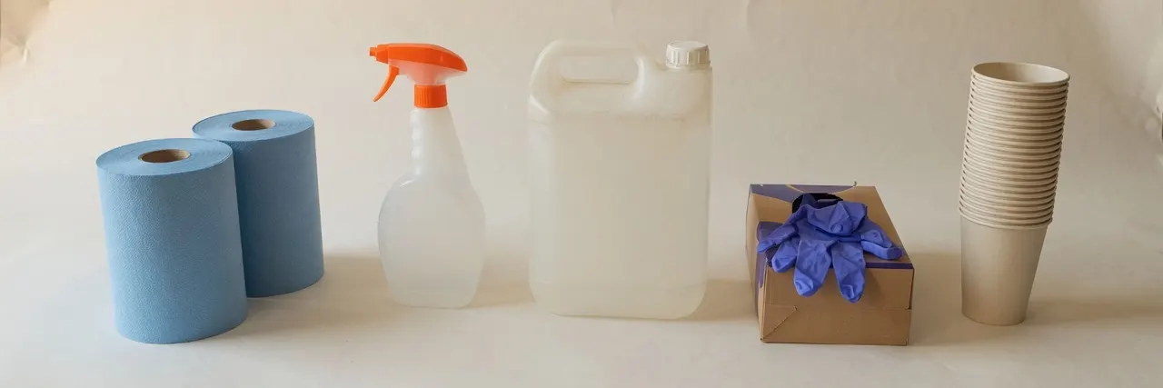 Rolls of blue paper towel, a spray bottle with an orange trigger, a 5 litre container, nitrile gloves and paper cups on warm paper